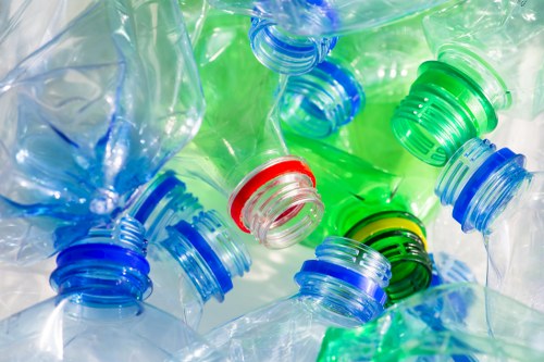 A close-up image showing several crushed plastic bottles with colourful caps, including blue, green, red, and black, scattered on a white surface. The bottles are made of transparent PET plastic and exhibit a glossy, crumpled texture indicating they are empty and discarded. The caps are partially screwed onto the bottles, with some caps lying loose nearby. The background features a softly blurred environment, emphasizing the focus on the plastic bottles and caps. This image visually relates to waste collection and rubbish removal services in the local area, such as those provided by House Clearance Egham, which handles the collection and proper disposal of household plastics and other rubbish types in the Runnymede or surrounding postcode areas near Egham.