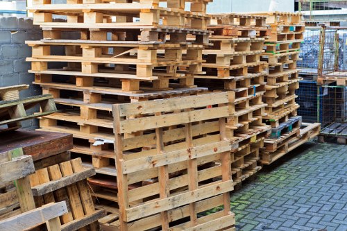 A large stack of new wooden pallets arranged outdoors on a paved surface, with some pallets leaning against the stack and others lying flat. The pallets are constructed from light-colored, untreated timber, showing a combination of horizontal and vertical planks with visible nails securing the joints. To the left, there are additional pallets and some weathered wooden crates, indicating an environment associated with warehouse or distribution storage. In the background, a metal fence and part of a building are partially visible, suggesting an industrial or commercial premises, possibly in Egham. The lighting is natural, highlighting the textures of the raw timber surfaces, which range from smooth to slightly rough with some knots and grain visible. The scene reflects a setting where rubbish removal services, such as those provided by House Clearance Egham, might collect or manage such pallet stacks as part of a house or business clearance process.