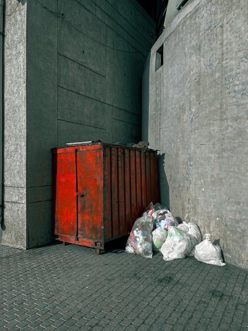 A pile of discarded cardboard boxes, some flattened and others still partially assembled, are placed on the ground against a weathered brick and concrete wall. The boxes vary in size and are made of b