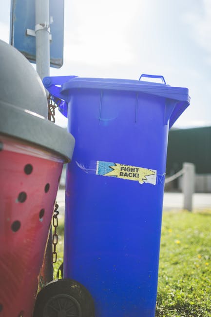 A tall, rectangular blue wheelie bin with a hinged lid is positioned outdoors on a paved surface. The bin features a bright yellow sticker with a blue border and the text 'FIGHT BACK!' printed on it.