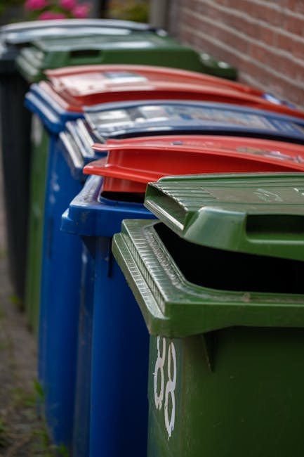 A large green waste disposal bin positioned on a paved surface next to a dark brick wall, filled with several red and black bags of garden or yard waste, including soil or mulch bags with visible bran